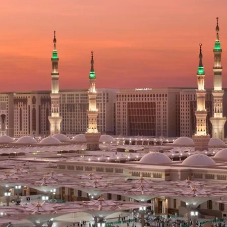 Готель Intercontinental Dar Al Iman Madinah By Ihg Medina Екстер'єр фото The photo depicts a scenic view of a mosque complex, likely during sunset, with several minarets illuminated and showcasing green lights. The architecture features domed roofs, and the setting includes a backdrop of buildings, all bathed in a warm, o