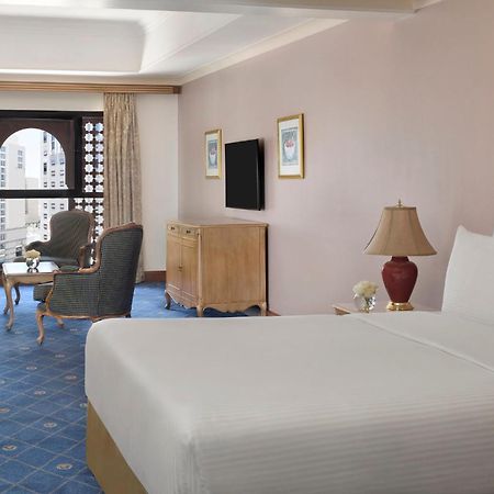 Готель Intercontinental Dar Al Iman Madinah By Ihg Medina Екстер'єр фото The photo shows a well-furnished hotel room. It features a large bed with a white duvet, two bedside lamps, and decorative paintings on the walls. There is a comfortable seating area with two chairs and a small table. A wooden dresser is positioned a
