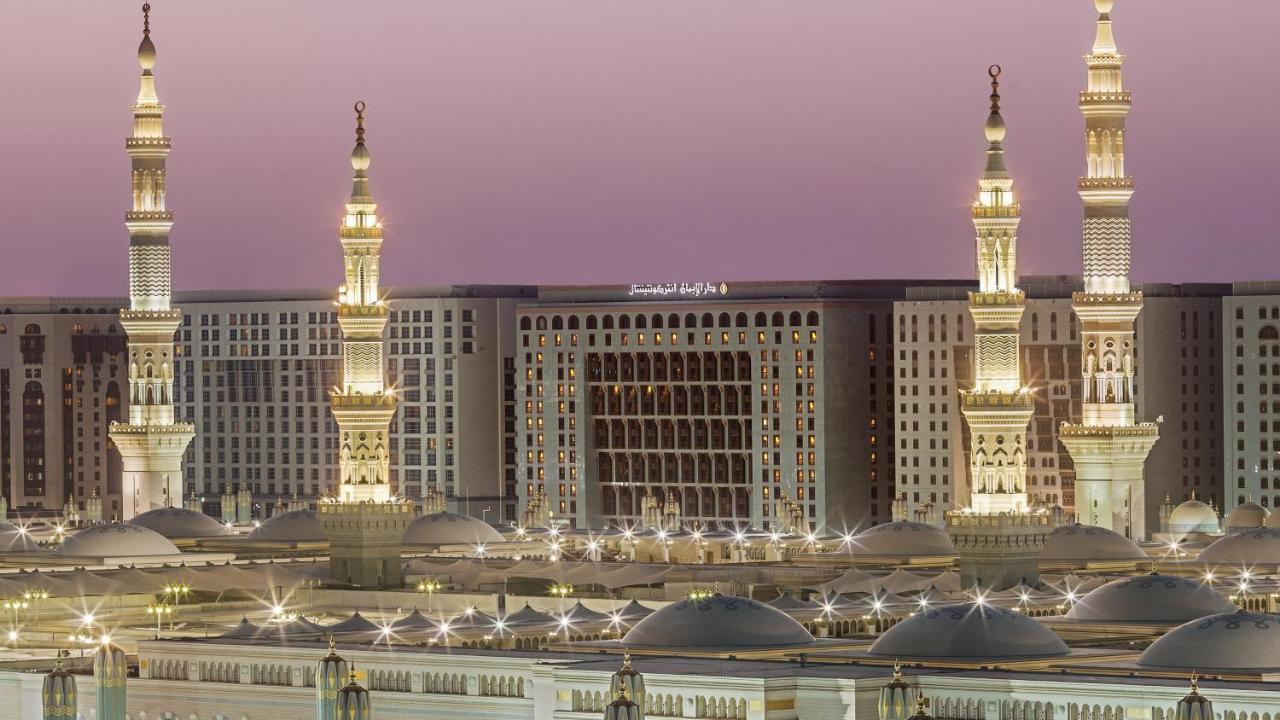 Готель Intercontinental Dar Al Iman Madinah By Ihg Medina Екстер'єр фото The photo depicts an architectural scene, likely from a mosque complex, featuring prominent minarets that are illuminated. The setting appears to be during twilight, with a soft gradient in the sky. In the background, a large building is visible, pos