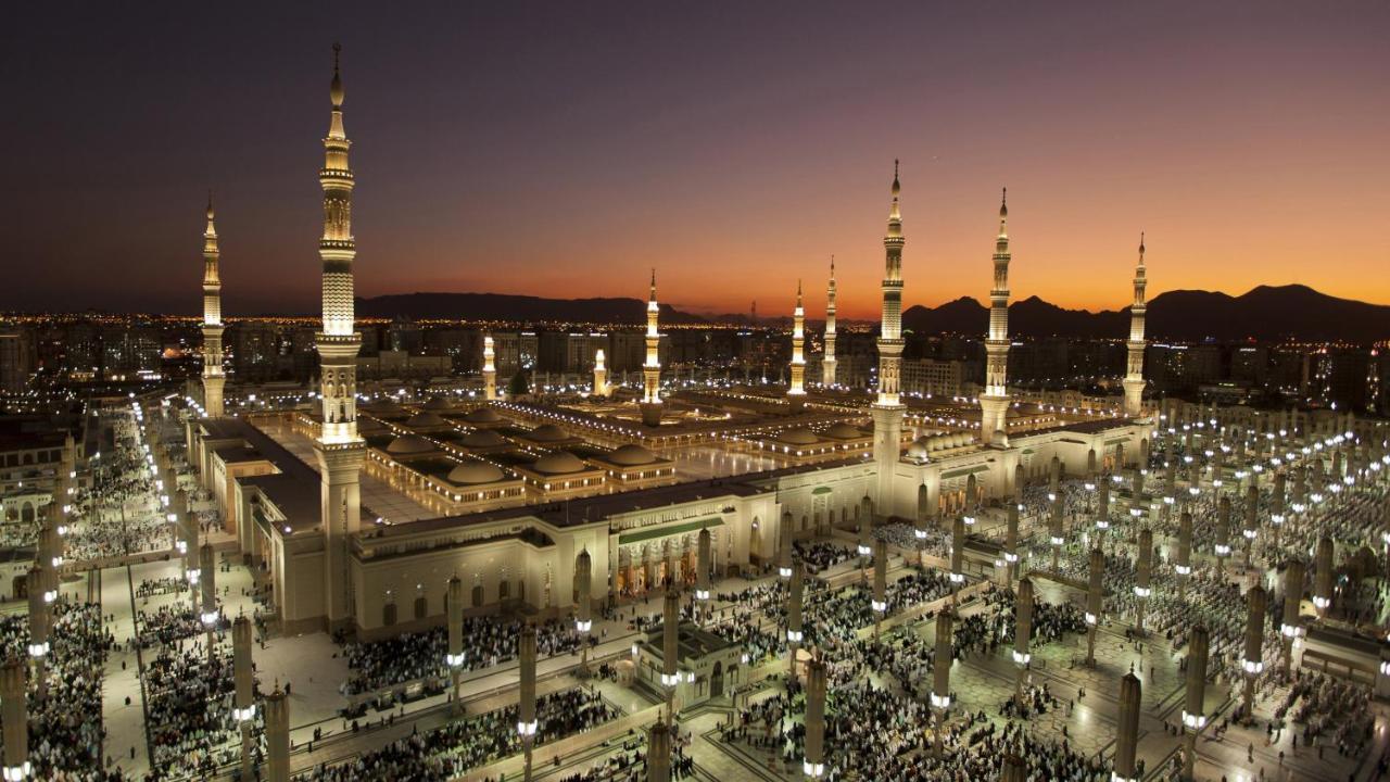 Готель Intercontinental Dar Al Iman Madinah By Ihg Medina Екстер'єр фото The photo depicts an aerial view of a large mosque complex during sunset. The mosque features multiple tall minarets, which are intricately designed and illuminated, giving the scene a stunning appearance as the sky transitions from daylight to dusk.