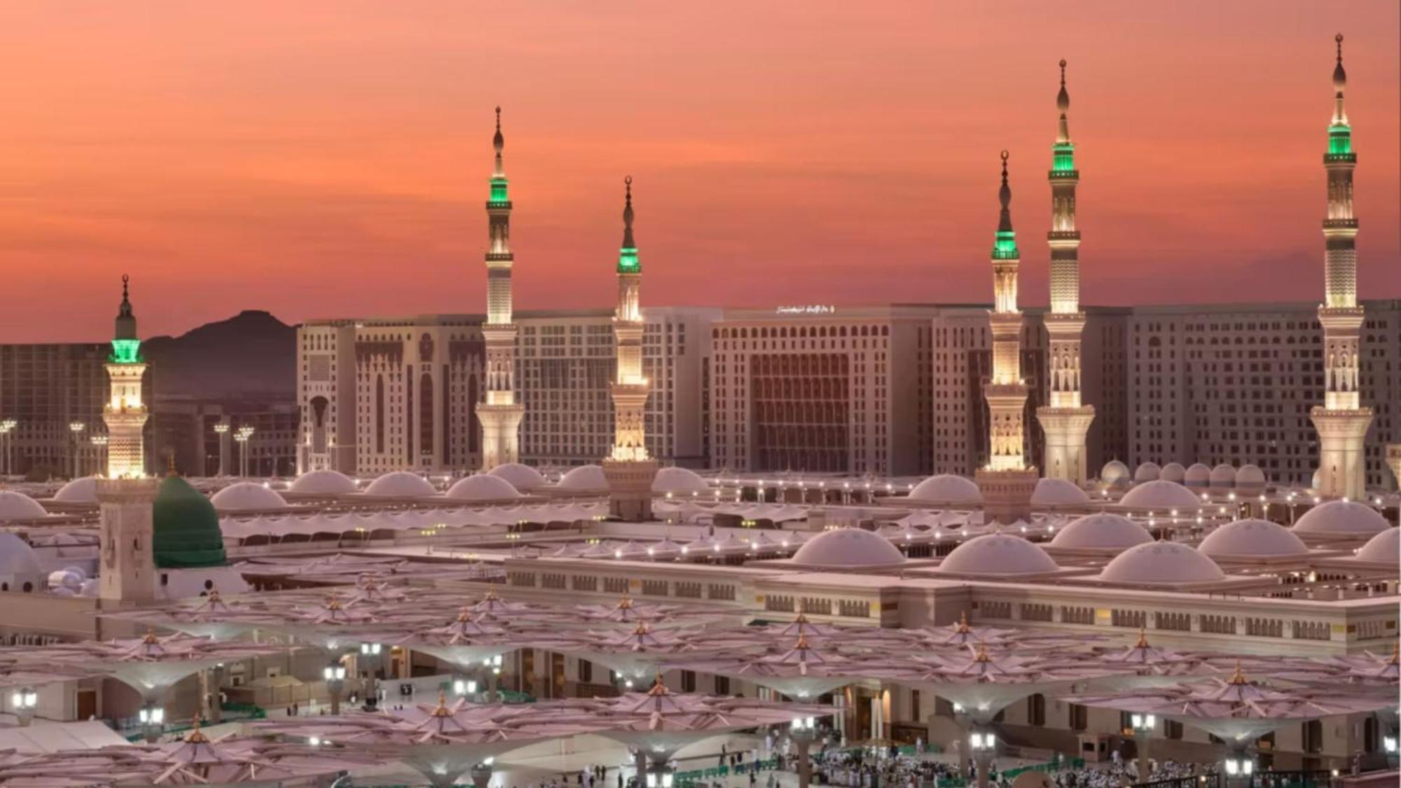 Готель Intercontinental Dar Al Iman Madinah By Ihg Medina Екстер'єр фото The photo depicts a scenic view of a mosque complex, likely during sunset, with several minarets illuminated and showcasing green lights. The architecture features domed roofs, and the setting includes a backdrop of buildings, all bathed in a warm, o