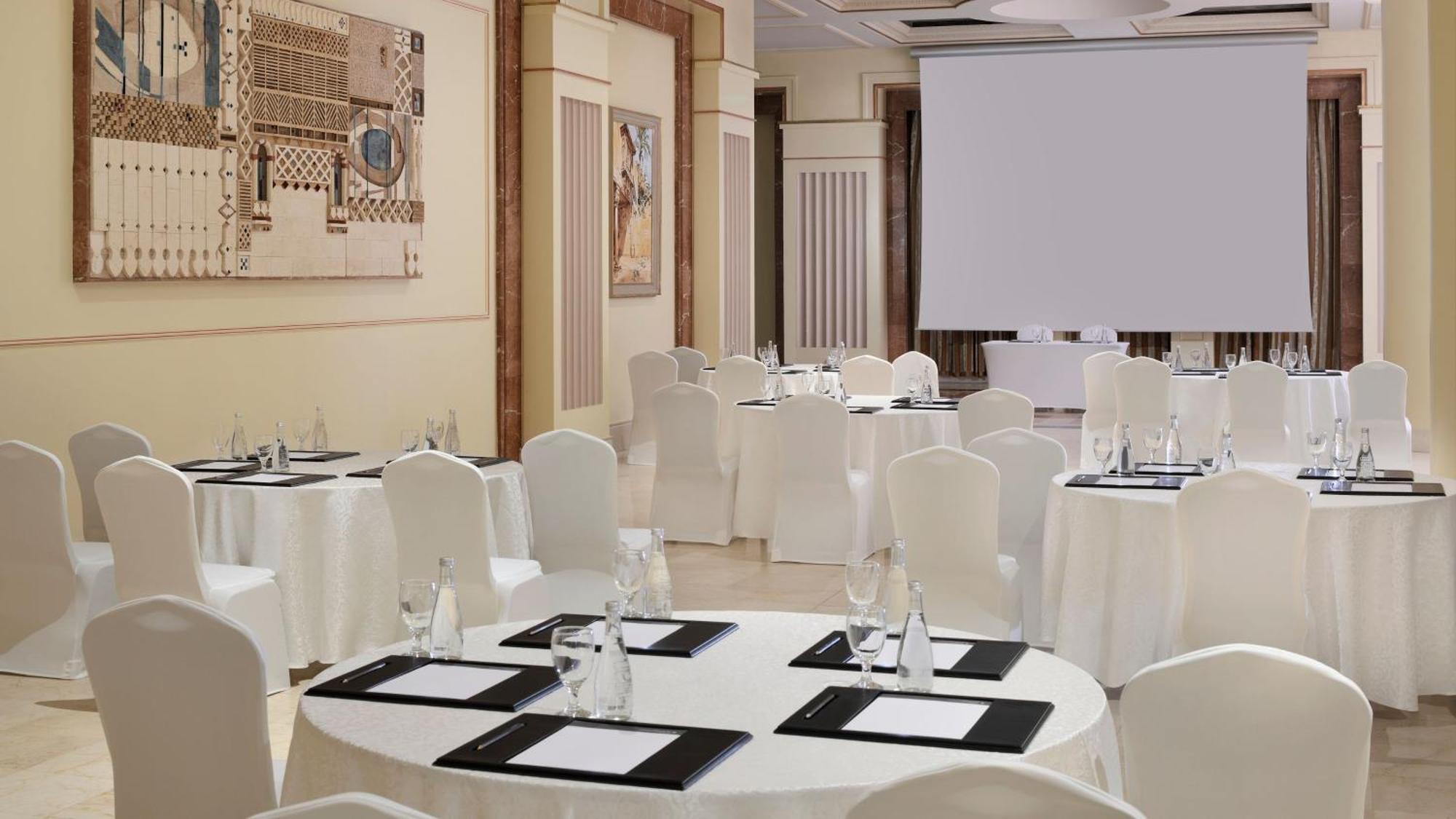 Готель Intercontinental Dar Al Iman Madinah By Ihg Medina Екстер'єр фото The photo depicts an elegantly arranged event space. There are several round tables covered with white tablecloths and surrounded by white chairs. Each table is set with a black folder or menu and a glass of water. The walls are adorned with artworks