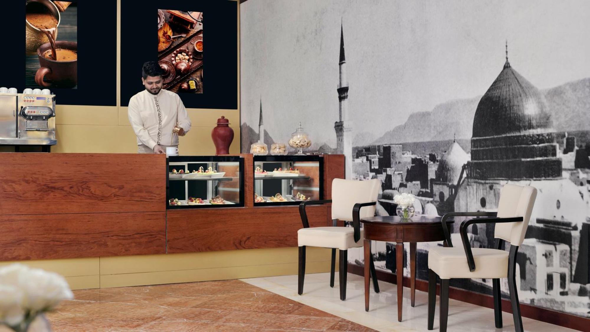 Готель Intercontinental Dar Al Iman Madinah By Ihg Medina Екстер'єр фото The photo shows an elegant cafe or dessert shop interior. A man in traditional attire is standing behind a wooden counter, where various pastries and sweets are displayed. The walls feature a large black-and-white mural of a cityscape, possibly depic