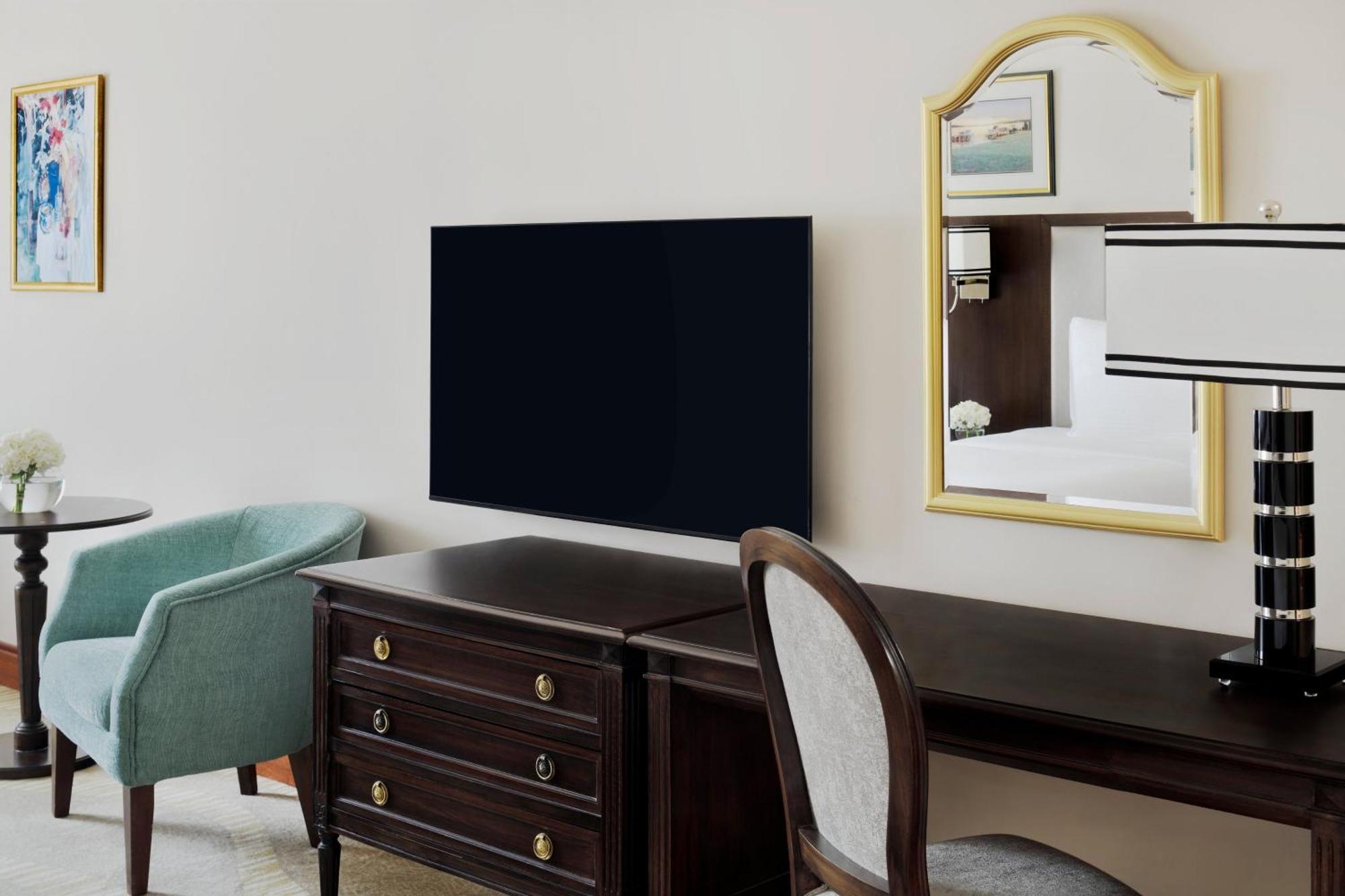 Готель Intercontinental Dar Al Iman Madinah By Ihg Medina Екстер'єр фото The photo shows a hotel room interior featuring a mounted flat-screen television on the wall. Below the TV, there is a dark wooden dresser with drawers. To the right, there's a wooden desk and a chair with a light-colored seat. A small table with a l