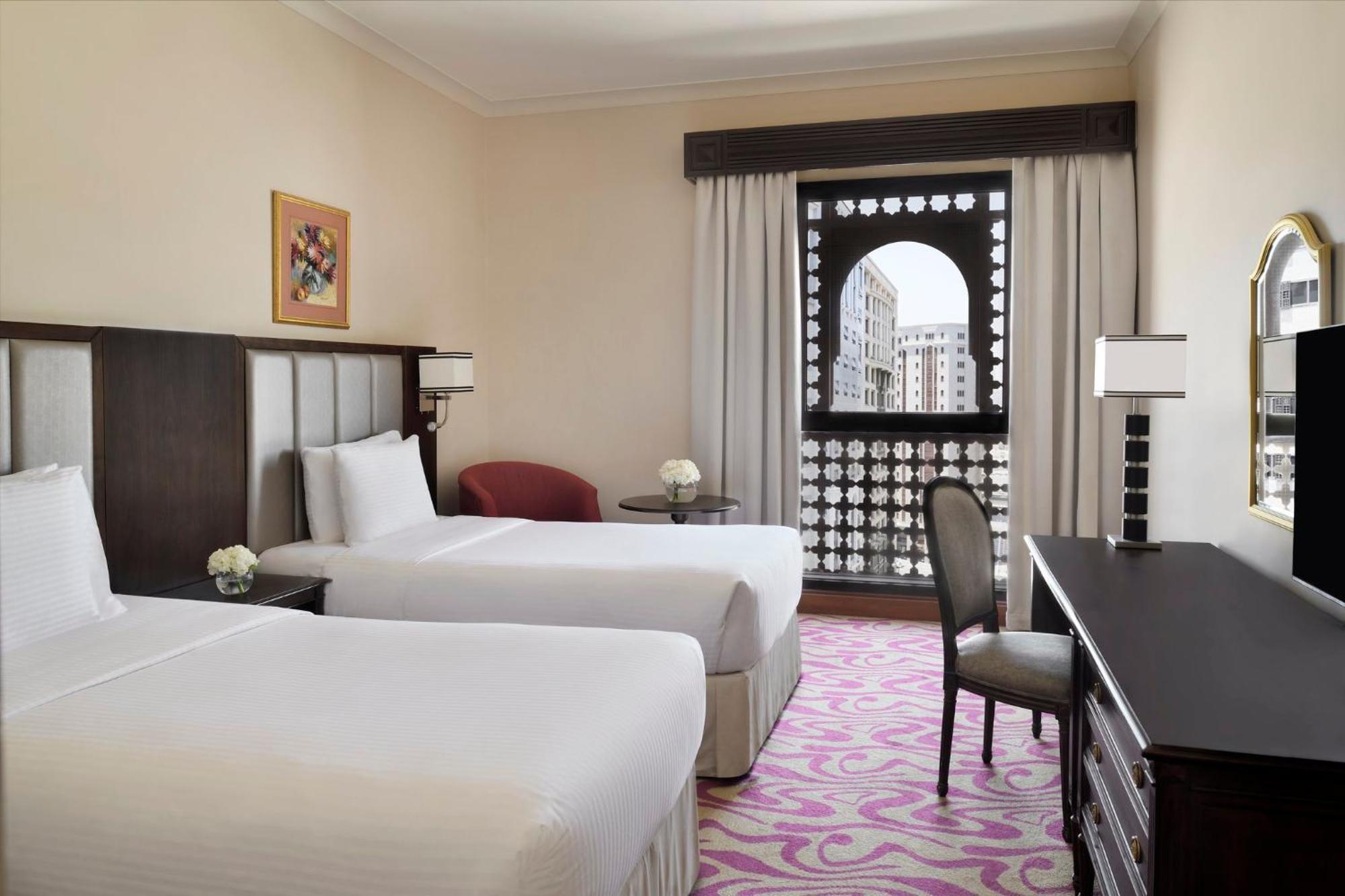 Готель Intercontinental Dar Al Iman Madinah By Ihg Medina Екстер'єр фото The photo shows a hotel room featuring two single beds with white bedding. The room has a light, neutral color scheme and is decorated with minimal furnishings. There's a small seating area with a red chair and a round table, as well as a desk with a