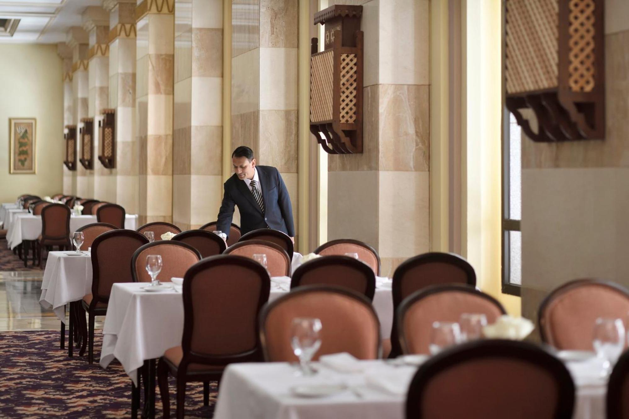 Готель Intercontinental Dar Al Iman Madinah By Ihg Medina Екстер'єр фото The photo depicts a dining area in an elegant restaurant. There are several tables set with white tablecloths, each accompanied by chairs. A waiter, dressed in a formal suit, is seen arranging or tending to one of the tables. The restaurant features 