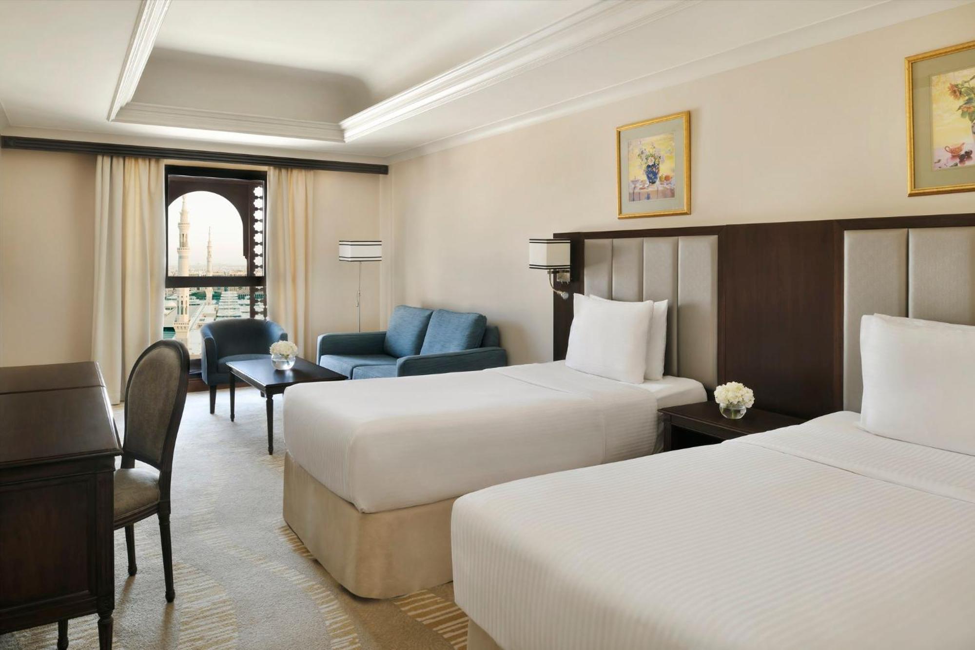 Готель Intercontinental Dar Al Iman Madinah By Ihg Medina Екстер'єр фото The photo shows a well-appointed hotel room featuring two twin beds with white bedding. There is a small desk with a chair, a comfortable blue sofa, and a coffee table in the sitting area. The room has a warm, neutral color scheme, accented by two pa