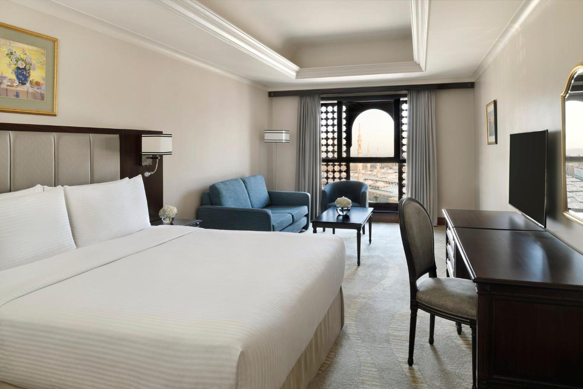 Готель Intercontinental Dar Al Iman Madinah By Ihg Medina Екстер'єр фото The photo shows a hotel room designed with a modern and comfortable aesthetic. It features a large bed with white linens, a seating area with a blue sofa, and a small table. There is a wooden desk with a chair and a television opposite the bed. Natur