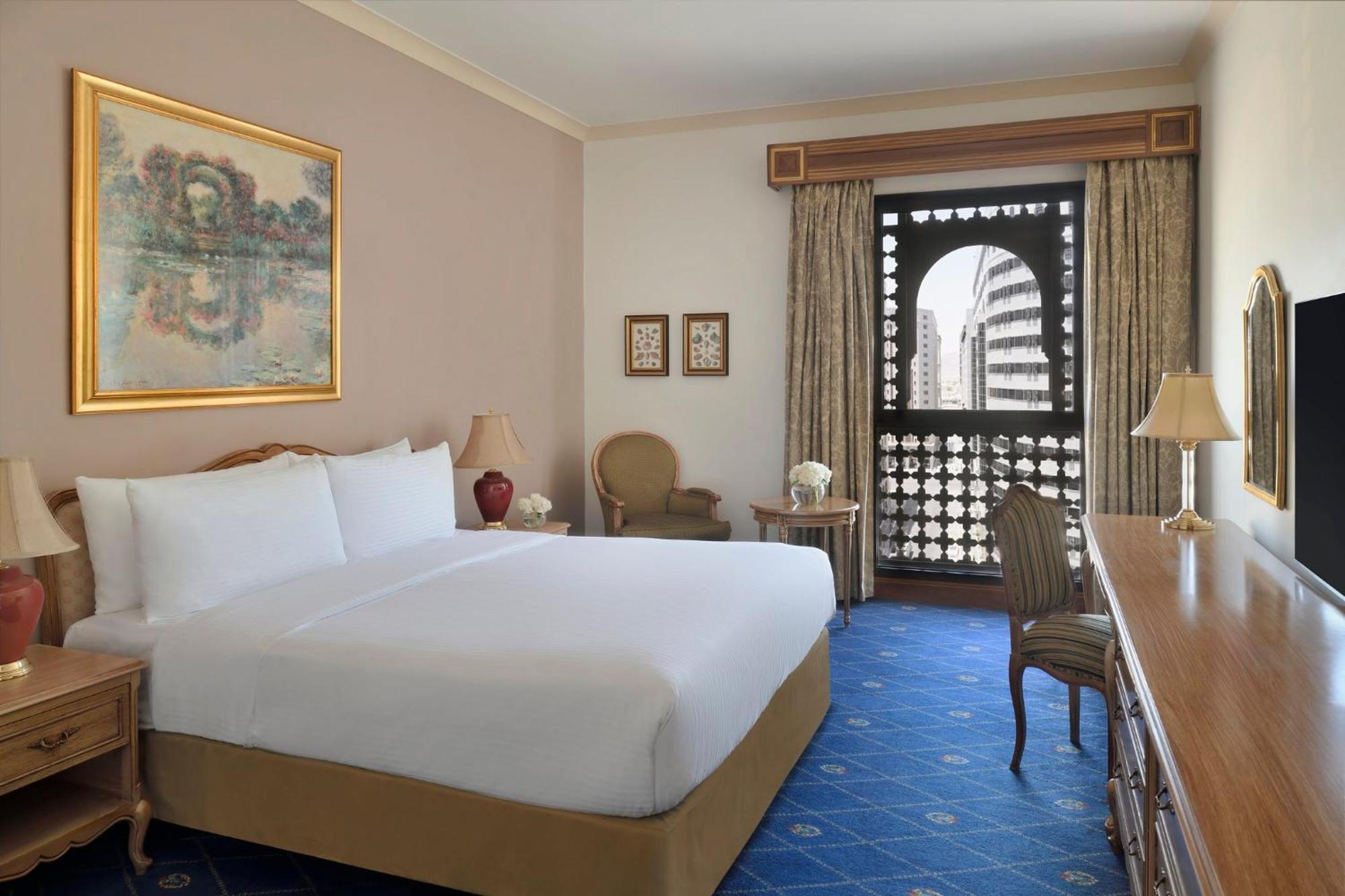 Готель Intercontinental Dar Al Iman Madinah By Ihg Medina Екстер'єр фото The photo shows a hotel room that features a large, neatly made bed with white linens. There is a framed painting on the wall above the bed. To the side, a small seating area includes a chair and a side table. Opposite the bed, there is a wooden desk
