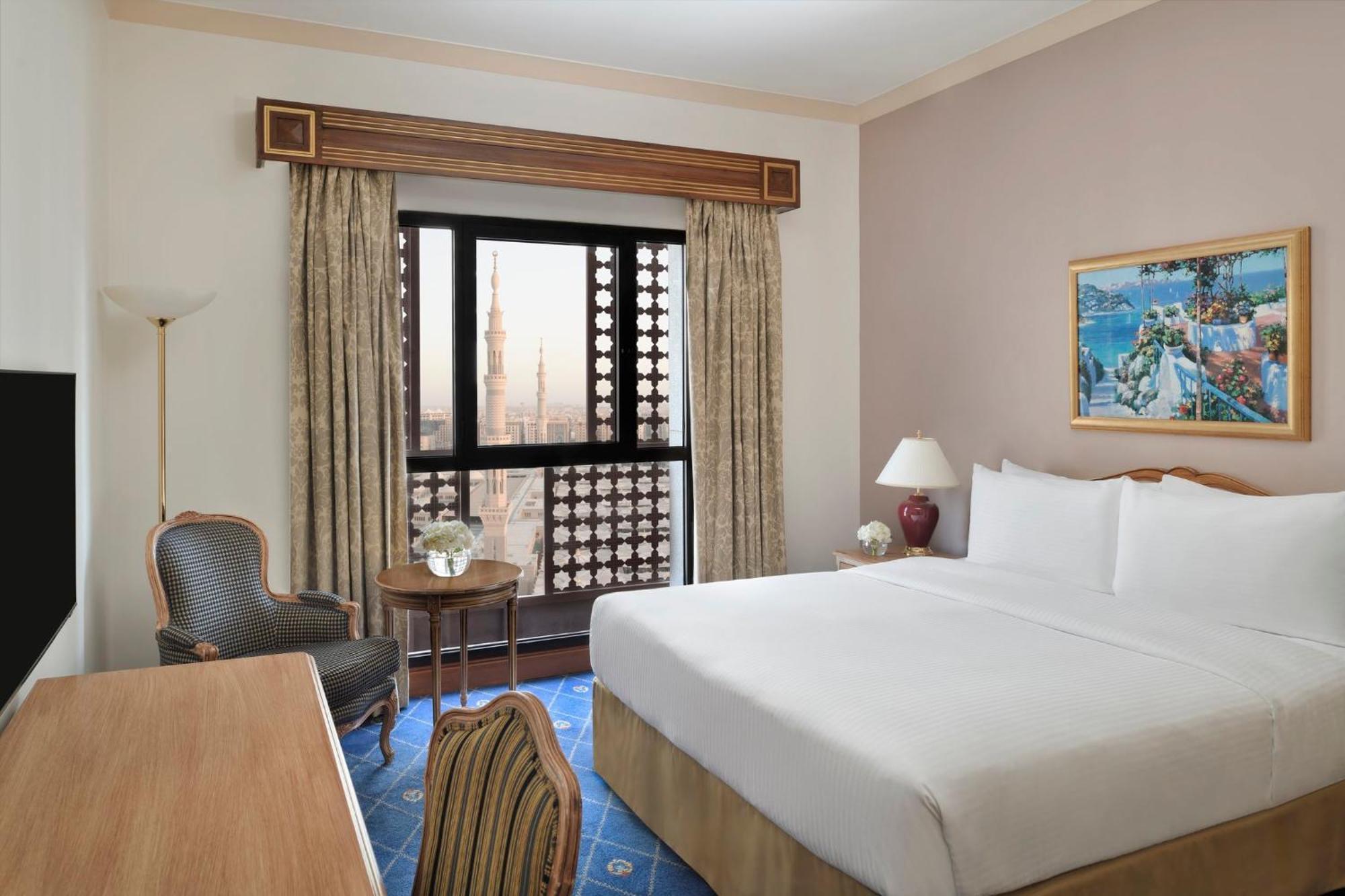 Готель Intercontinental Dar Al Iman Madinah By Ihg Medina Екстер'єр фото The photo shows a hotel room with a modern design. There is a large bed with white linens placed against the wall. Next to the bed, there is a small bedside table with a lamp. A comfortable armchair is situated nearby, accompanied by a wooden desk. T