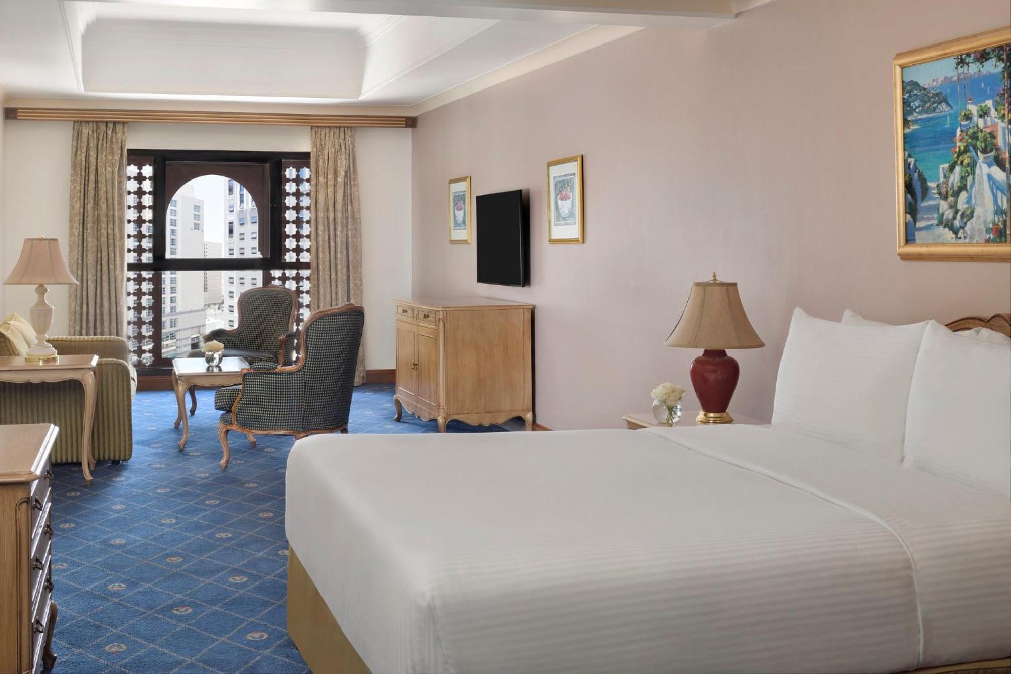 Готель Intercontinental Dar Al Iman Madinah By Ihg Medina Екстер'єр фото The photo shows a well-furnished hotel room. It features a large bed with a white duvet, two bedside lamps, and decorative paintings on the walls. There is a comfortable seating area with two chairs and a small table. A wooden dresser is positioned a