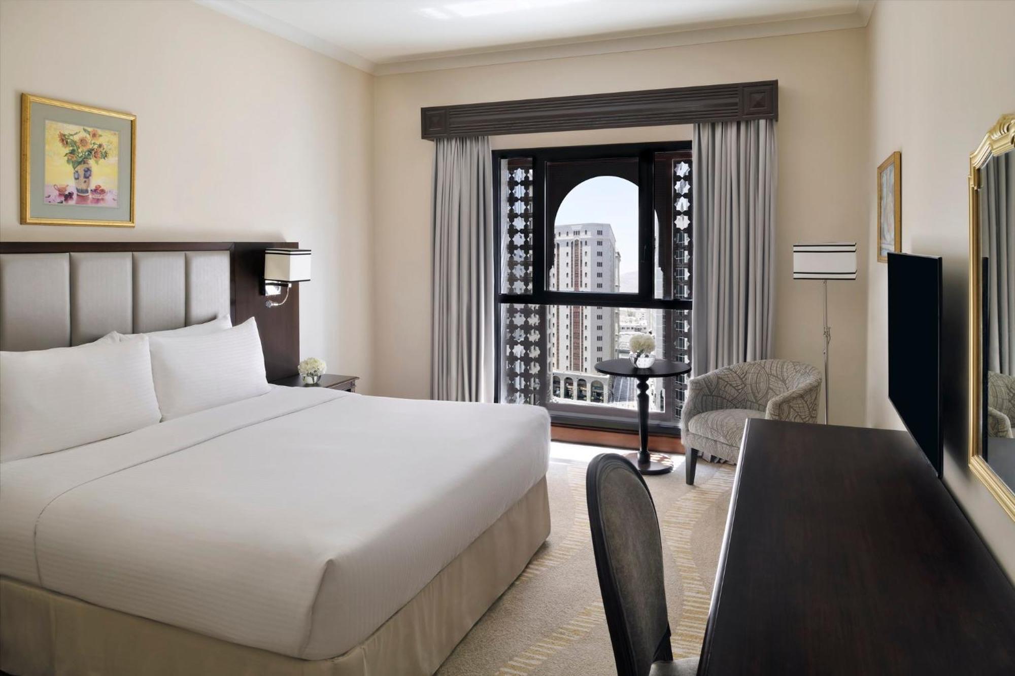 Готель Intercontinental Dar Al Iman Madinah By Ihg Medina Екстер'єр фото The photo shows a hotel room with a modern and minimalistic design. It features a large bed with white linens, a side table, and a comfortable chair near a window. The window has a view of a cityscape, and elegant curtains frame the window. There is 