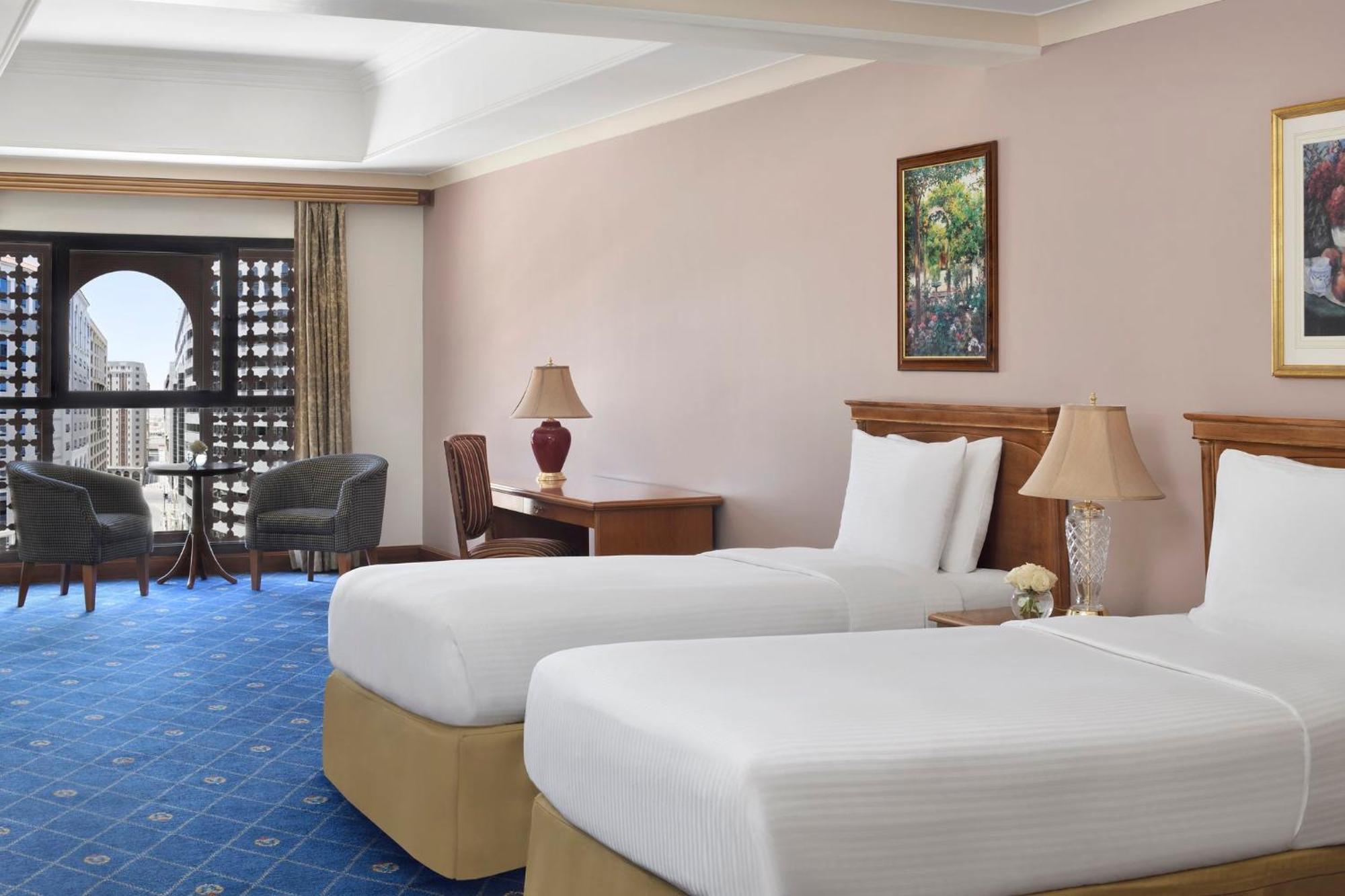 Готель Intercontinental Dar Al Iman Madinah By Ihg Medina Екстер'єр фото The photo shows a nicely designed hotel room featuring two twin beds with white linens and beige bases. In the background, there's a wooden desk and a lamp, along with a pair of comfortable armchairs positioned near a large window. The window showcas