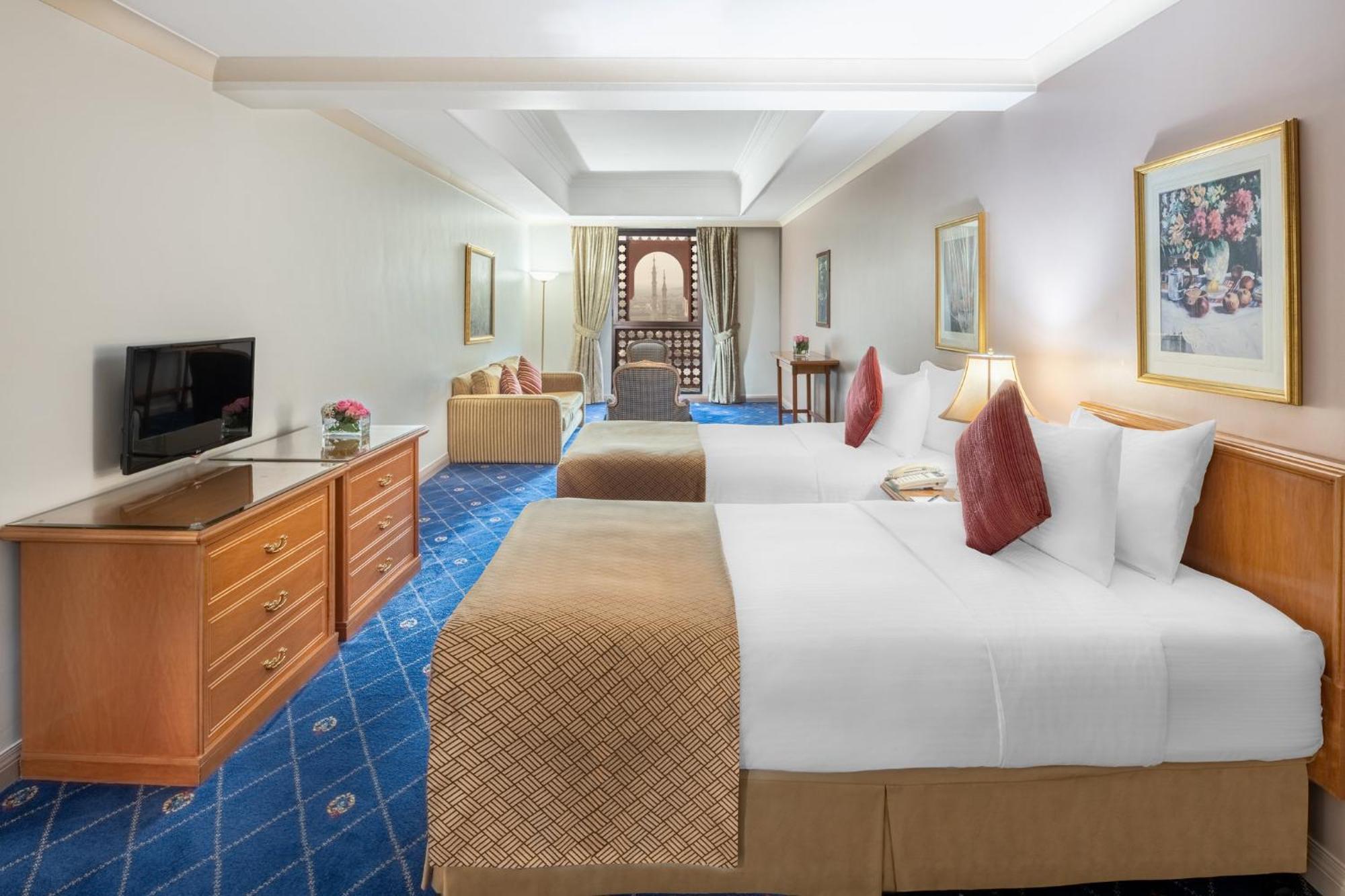 Готель Intercontinental Dar Al Iman Madinah By Ihg Medina Екстер'єр фото The photo shows a hotel room featuring two beds with white linens and decorative pillows. There is a wooden dresser with a television on it. The flooring has a blue carpet with a pattern, and the walls are painted in a light color. The room is well-l