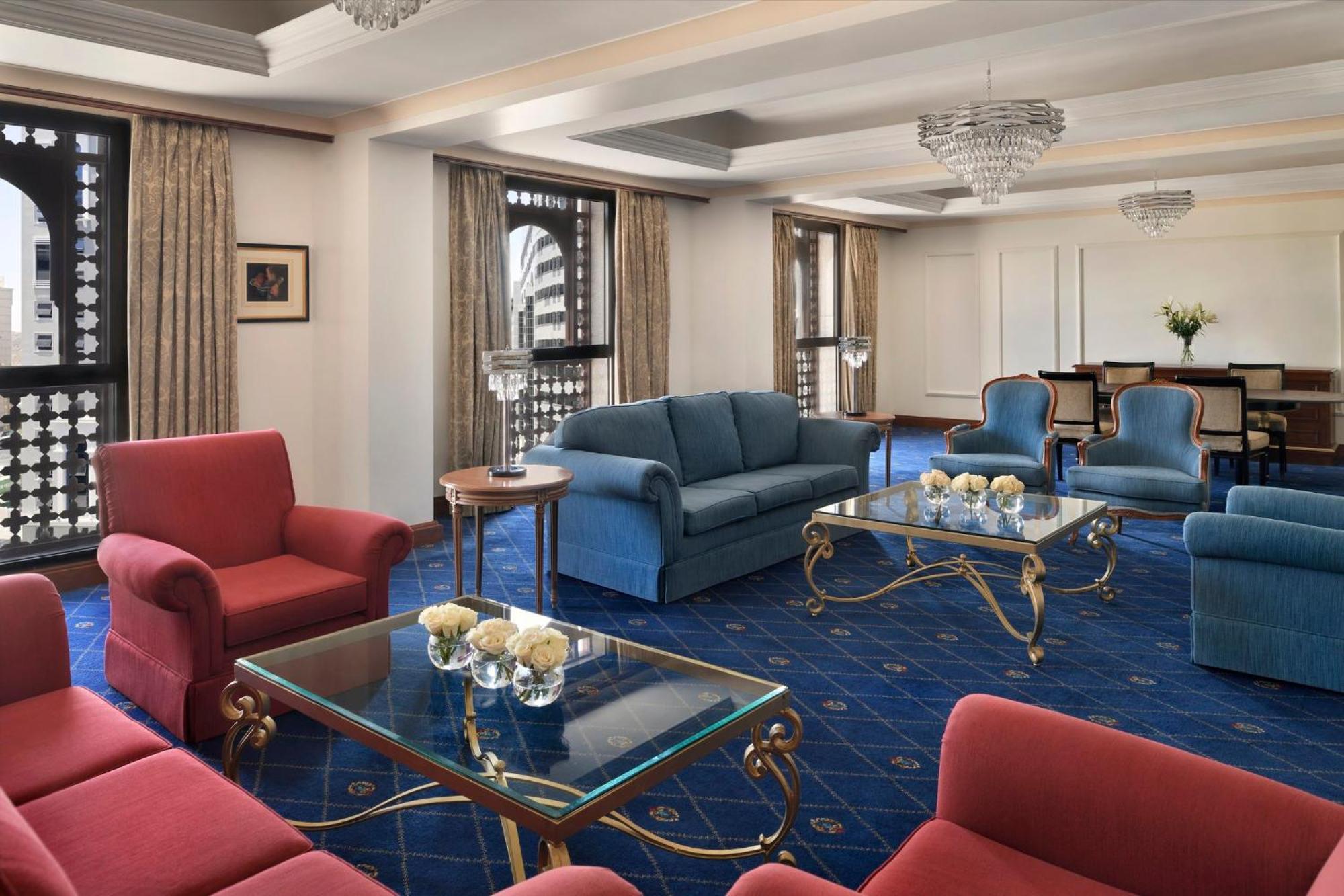 Готель Intercontinental Dar Al Iman Madinah By Ihg Medina Екстер'єр фото The photo shows a luxurious living space or lounge area. It features a combination of furniture, including a blue sofa, several red armchairs, and glass-top coffee tables with gold accents. The flooring is carpeted in a deep blue, and the walls are a