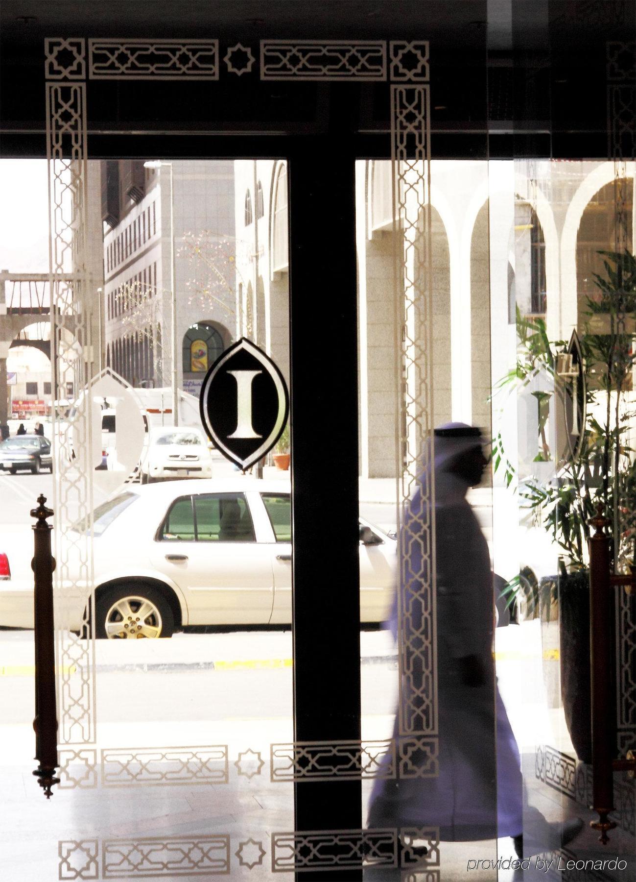 Готель Intercontinental Dar Al Iman Madinah By Ihg Medina Інтер'єр фото The photo shows a view through a glass door or window, featuring an elegant design with a prominent "I" symbol displayed. Through the glass, there are some parked vehicles visible, including a white car. In the foreground, a shadowy figure of a perso