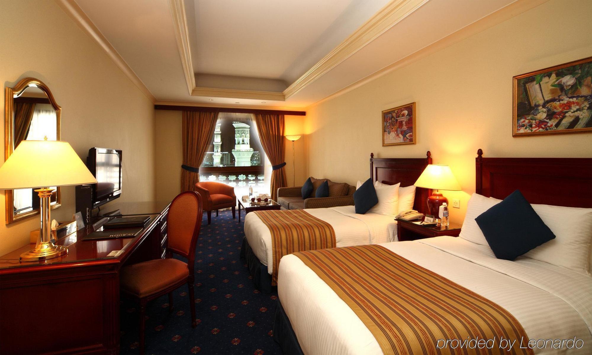 Готель Intercontinental Dar Al Iman Madinah By Ihg Medina Номер фото The photo shows a hotel room featuring two beds with white linens and brown striped throws. There is a wooden desk and chair, alongside a television. The walls are painted in a warm tone, and there are decorative paintings. Large windows with curtain