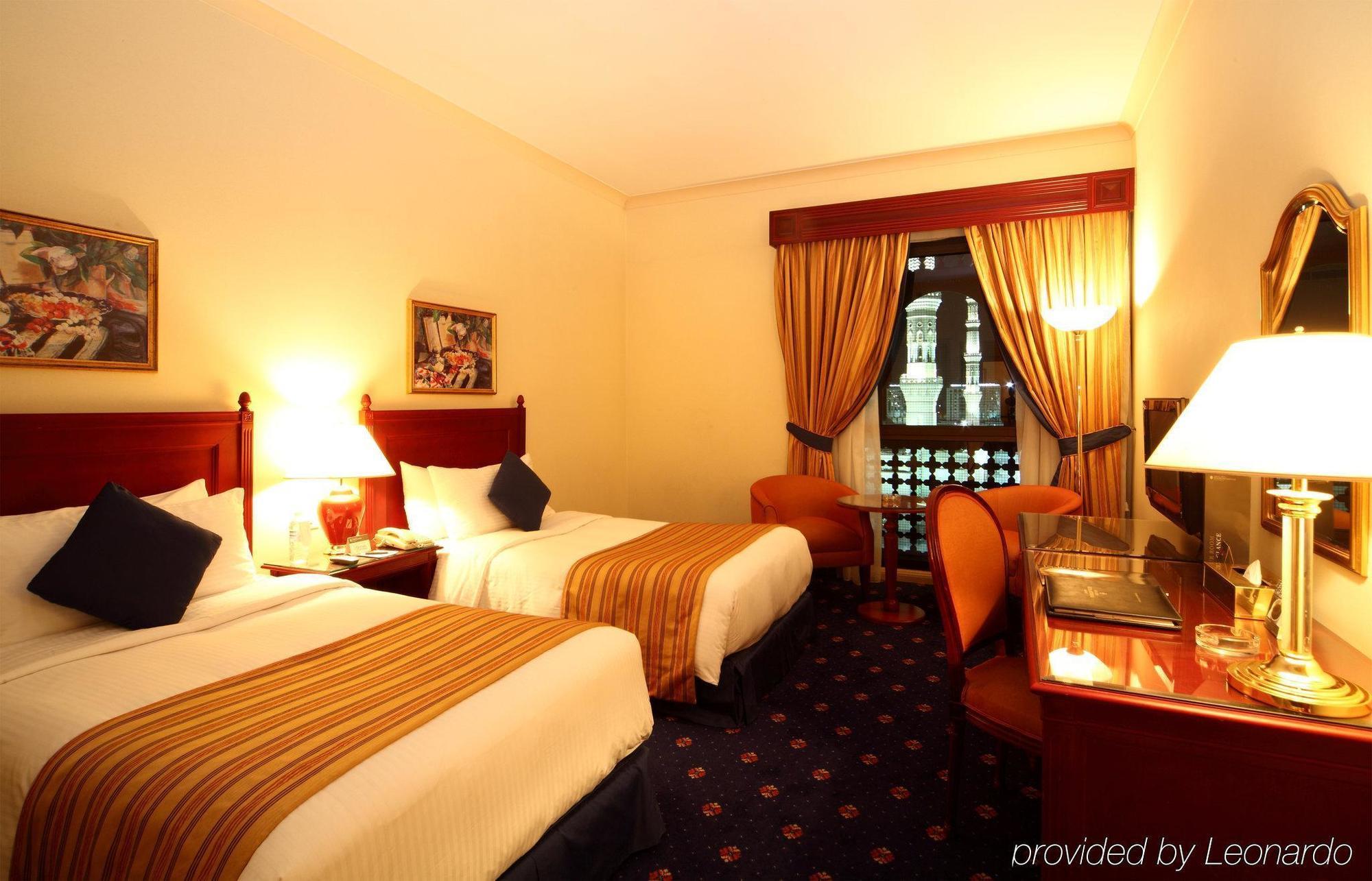 Готель Intercontinental Dar Al Iman Madinah By Ihg Medina Номер фото The photo shows a hotel room featuring two twin beds with white linens and brown striped bedspreads. There are decorative pillows on the beds, and the room has warm-colored walls. To the side, there's a small sitting area with a round table and a cha