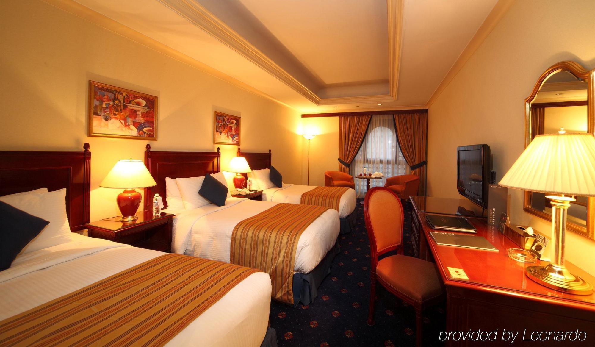 Готель Intercontinental Dar Al Iman Madinah By Ihg Medina Номер фото The photo shows a hotel room with a warm and inviting atmosphere. There are two beds with white and gold bedding, complemented by decorative pillows. Each bed has a bedside lamp. In the background, there's a small seating area with a couple of chairs