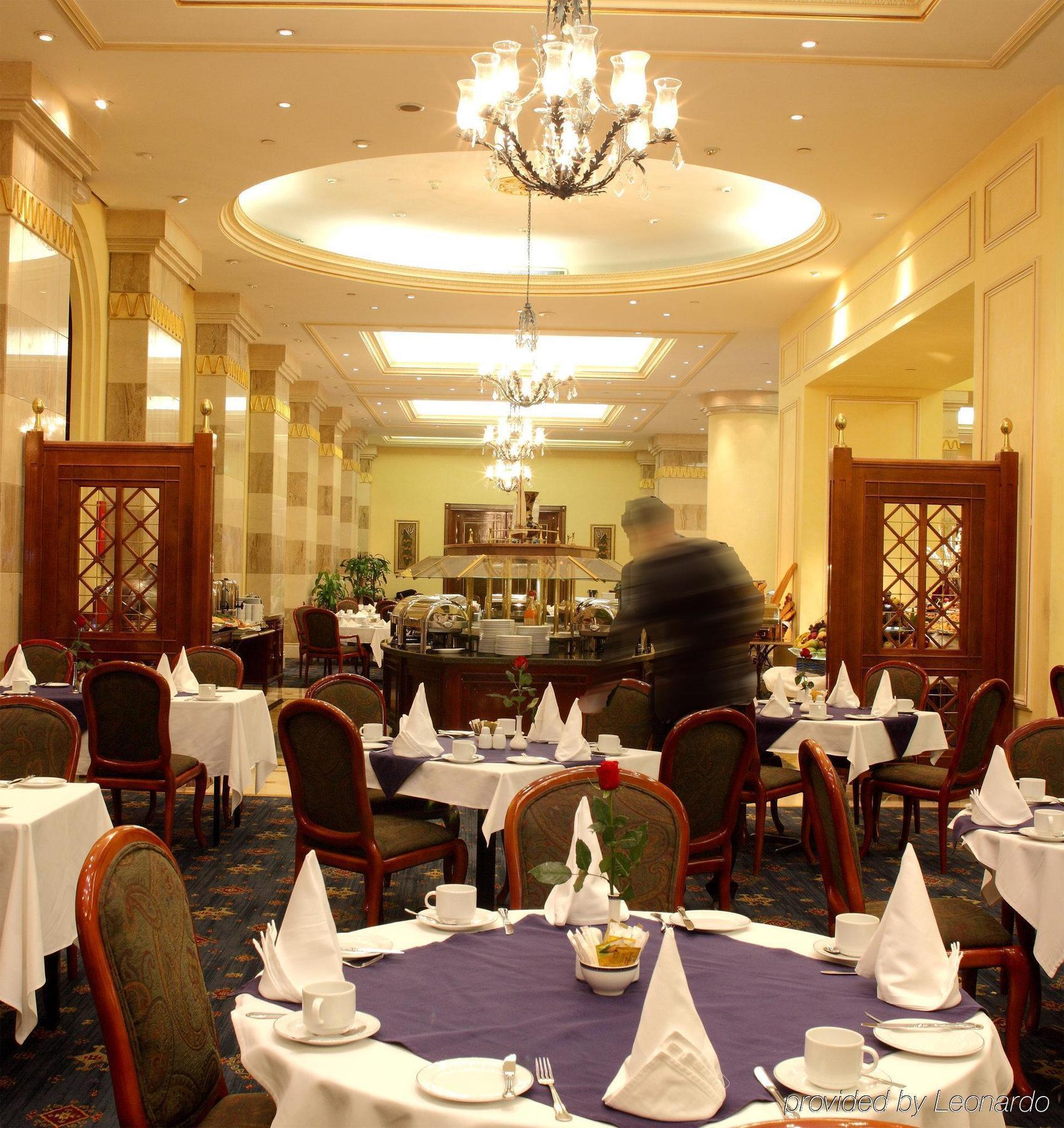 Готель Intercontinental Dar Al Iman Madinah By Ihg Medina Ресторан фото The photo depicts an elegant restaurant interior. It features round tables covered with white tablecloths, each set with plates, cutlery, and folded napkins arranged into triangles. The space has a warm, inviting atmosphere with a mix of decorative e