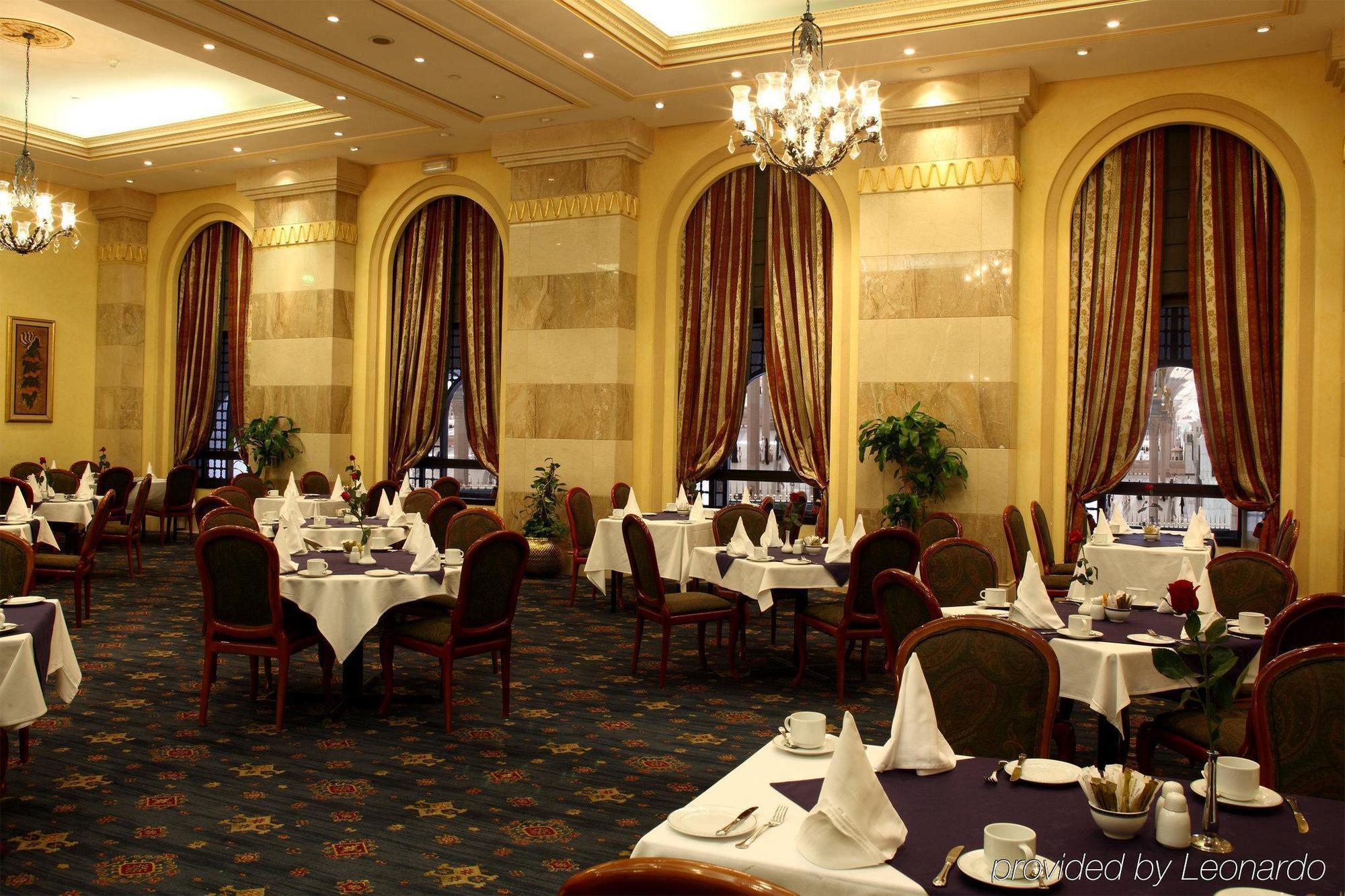 Готель Intercontinental Dar Al Iman Madinah By Ihg Medina Ресторан фото The photo depicts an elegant restaurant interior. The space features ornate decor with tall windows draped in rich, patterned curtains. Tables are neatly set with white tablecloths and napkins, and there are plush chairs arranged around them. The flo