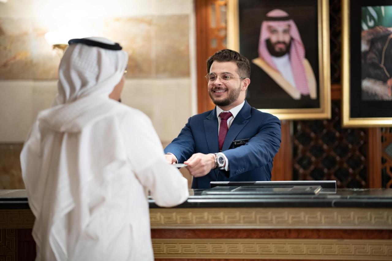 Готель Intercontinental Dar Al Iman Madinah By Ihg Medina Екстер'єр фото The photo shows a hotel reception area. A male receptionist, dressed in a navy suit with a tie, is smiling and handing an item to a guest who is wearing traditional white attire, possibly a kandura or thobe. The background features elegantly decorate